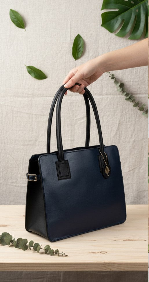 Navy blue and black plant leather tote bag held by hand on a wooden surface with green leaves and a neutral fabric background